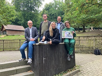 Drei Männer und zwei Frauen stehen und sitzen vor einem Gehege. Eine Frau hält einen Kalender in der Hand.